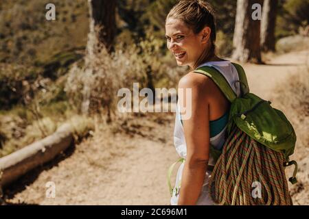 Woman with climbing rope in backpack hiking up a mountain. Female mountaineer looking back at the camera and smiling while walking on mountain trail. Stock Photo