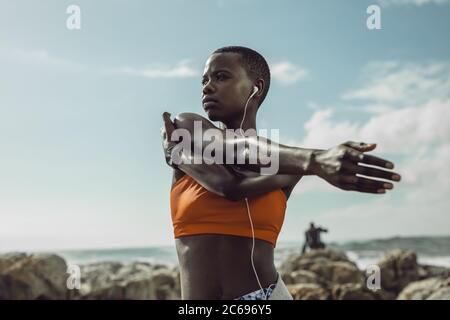 African female with headphones stretching her arms outdoors. Woman in sportswear exercising at the beach. Stock Photo