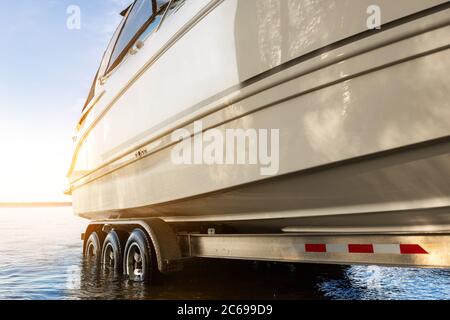 Big luxury cabin motorboat cruiser yacht launching at trailer ramp on river or lake. Warm morning sunrise sunshine reflection in calm water surface Stock Photo