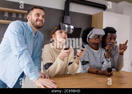 Multicultural group of friends playing video games wearing virtual reality glasses with controllers, women trying to play and boyfriends making hints Stock Photo