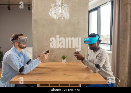 Two male entrepreneurs in casual clothing, sitting opposite each other at table in empty room, being busy with developing a project using virtual real Stock Photo