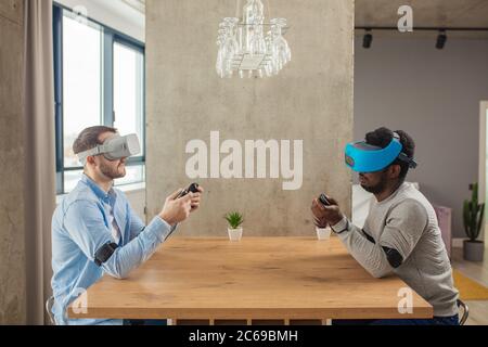 Cheerful interracial male gamers having fun with new trends technology, sitting against each other at table with virtual reality headset on . Stock Photo
