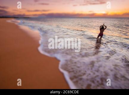 A blurred image of a woman (MR) standing in the surf, looking out at the sunset on the two mile long, three hundred feet wide, Papohaku Beach on the w