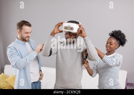 Photo of happy african american couple and caucasian young man standing in room and laughing while using 3d virtual reality device. Stock Photo