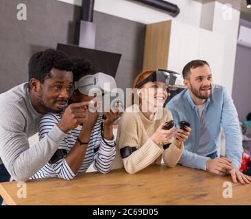 Multicultural group of friends playing video games wearing virtual reality glasses with controllers, women trying to play and boyfriends making hints Stock Photo
