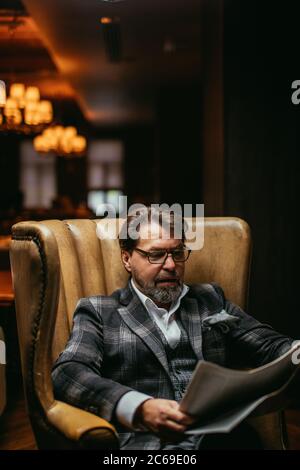 Handsome middle-aged prosperous businessman sitting in leather arm-chair in hotel lobby, wears optical spectacles is reading newspaper while waiting f Stock Photo