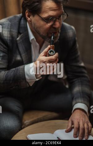 Concentrated mature start-up project developer making notes on the paper wordpad, smoking pipe with contemplating expression, sitting in the lobby of Stock Photo