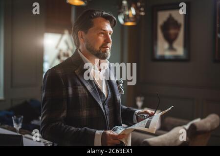 Confident senior business man in elegant custom made suit reading fresh newspaper while standing in restaurant or cafe Stock Photo