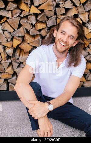 Portrait of a handsome man sitting on ground leaning against a log pile, Russia Stock Photo