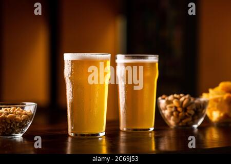 Two glasses of lager on wooden table, near are pistachios, nuts and chips in glass plates Stock Photo