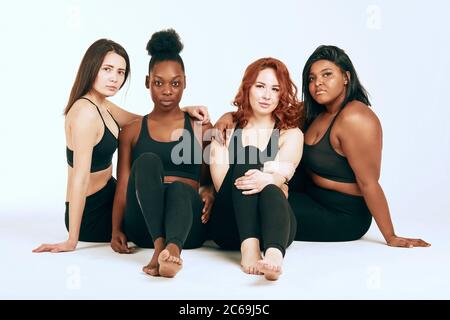 Group of women of different race, figure and size in sportswear sitting together as group, looking confidently at camera against white background. Stock Photo