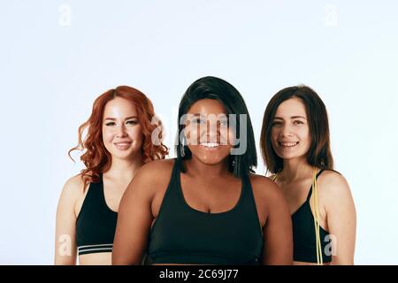 African happy overweight woman in company of two caucasian slim girlfriends, dressed in sportive clothes . Fitness, sport, diet concept. Stock Photo