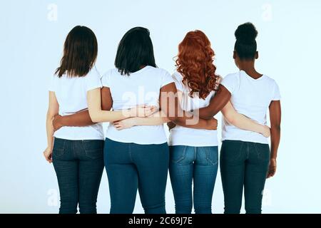 Rear view of diverse multi ethnic women with different body size and hairstyle standing in row dressed in the same casual manner, hugging together on Stock Photo