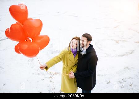 happy caucasian couple, young man and woman spend romantic pastime together, walking at winter street with red air balloons, celebrating their anniver Stock Photo