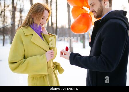 romantic caucasian bearded man in coat making marriage proposal to his girlfriend at winter street. man hold small red flocked ring box and red air ba Stock Photo