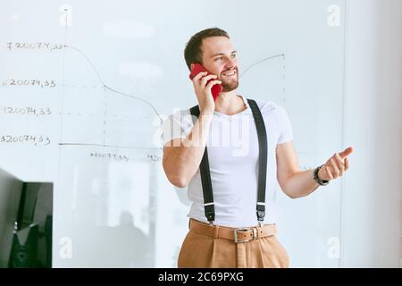 Young businessman wearing white t-shirt and trousers with suspenders look side, talking on phone with business partners. Flipchart diagrams background Stock Photo
