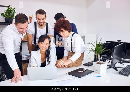 young and enthusiastic people in office, stand near asian woman sitting with laptop. Discuss new project looking at screen of laptop, smiling Stock Photo