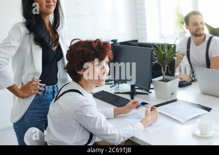 Beautiful smiling woman look side at coach, attentively listen to speaking,colleagues around her, sit on table with computers, papers Stock Photo