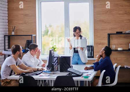 Young multi-ethnic team of business partners sit listen to successful speaking of experienced asian woman holding white laptop. African, caucasian and Stock Photo
