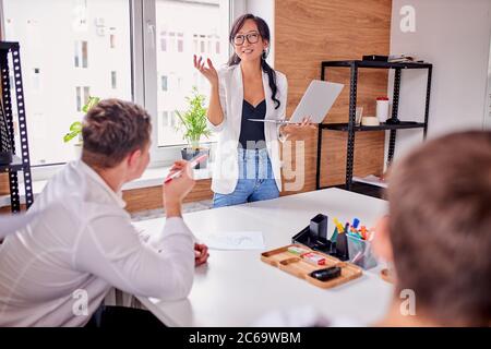 Smiling asian business woman in white shirt and glasses look at listeners and laugh, isolated in office. stand while others sitting Stock Photo