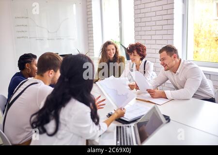 Friendly and cheerful colleagues in meeting office, sit on table having conversation, holding papers Stock Photo