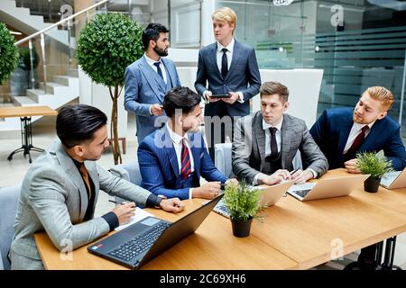 Young men gathered to discuss new project, use laptop, sit on table, young businessmen in office wearing tuxedo Stock Photo
