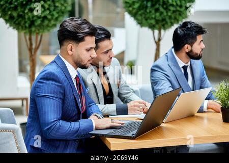 Side view on young politicians conduct negotiations about new project, new deal, using laptops, sitting on wooden table. Office background Stock Photo