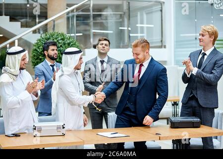 Sheikhs making deal with caucasian man in suit. Two businessmen shaking hands to each other. Witnesses applaud. Business center background Stock Photo