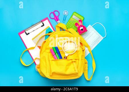 Back to school, education concept Yellow backpack with school supplies, protective medical mask, calculator, scissors isolated on blue background. Top Stock Photo