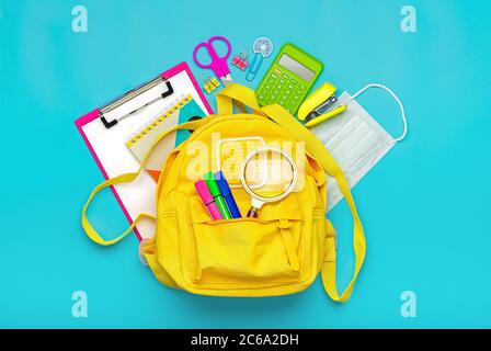 Back to school, education concept Yellow backpack with school supplies, protective medical mask, calculator, scissors isolated on blue background. Top Stock Photo