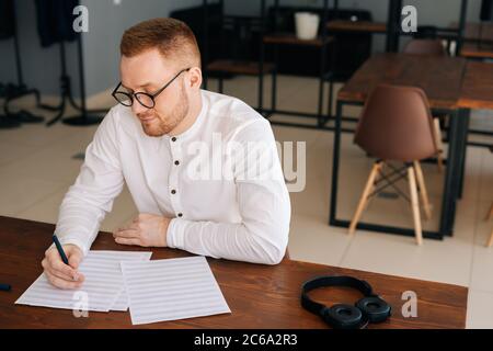Talented young composer wearing stylish eyeglasses writing musical notes with pen on sheet music Stock Photo