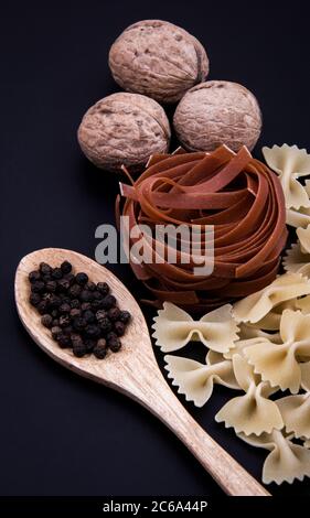 Colored and macaroni-shaped pasta in different sizes and colors with a bowl of black pepper and a few walnut kernels.... Stock Photo