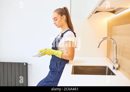 Side view on beautiful caucasian janitor in blue uniform stand wiping dishes in kitchen, white interior of kitchen. Young cleaner wearing yellow rubbe Stock Photo
