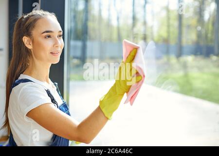 Cleaning window in cottage with panoramic window. Side view on caucasian woman in uniform, smiling. Panoramic window view on garden