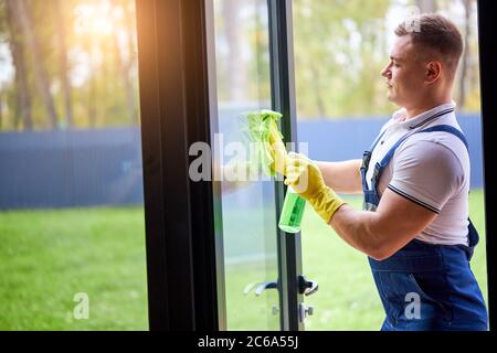 Young muscled guy in uniform from cleaning service wearing yellow protective gloves, holding spray wash the window Stock Photo