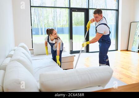 Two janitors in uniform cleaning new room with white sofa, mopping floor, straighten sofa Stock Photo