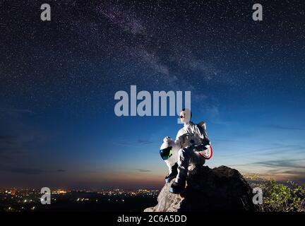 Side view of astronaut sitting on top of rocky hill and looking at beautiful night sky. Spaceman wearing space suit and holding helmet. Concept of astronautics and human space exploration. Stock Photo