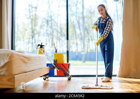 Process of mopping floor by young female in blue uniform, trolley of detergents next to her Stock Photo