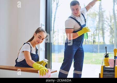 Side view on active job of two caucasian janitors dressed in uniform for cleaning, man clean glass of window, woman wiping dust off sofa. White interi Stock Photo