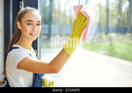 Careful young cleaner in yellow rubber gloves cleaning window with spray and rag. Cleaning service concept. Janitor lady look at camera Stock Photo