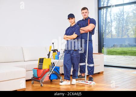 Two young members of cleaning service busy with house cleaning session, stand closely against living room background. Hygiene concept. Stock Photo