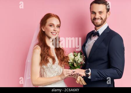 Happy male in suit and female in white wedding dress putting golden ring with diamond on finger, stand isolated over pink background. Love, marriage c Stock Photo