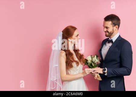 Lovely caucasian coule of young woman and man in wedding wear posing at camera isolated over pink background, look at each other Stock Photo