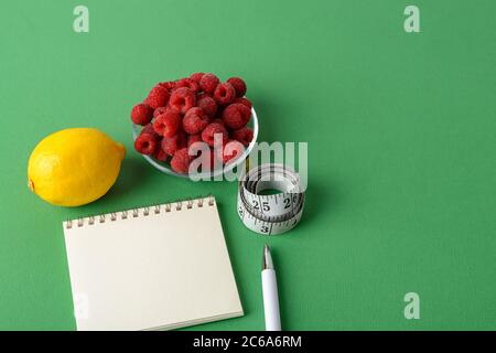 Inch measuring tape roll, lemon, raspberries, paper notepad and white pen on a green background. Healthy eating, diet, weight loss and body slim. Stock Photo