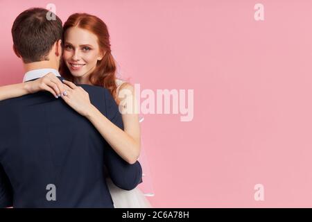 Rear view on man in tux with hugging him smiling woman, isolated over pink background. Love Stock Photo