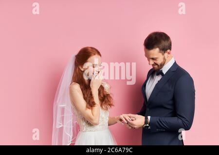 caucasian gentleman in tux with bow putting on gold diamond ring nd on finger to joyful cheerful bride with jewelry holding white wedding bouquet in h Stock Photo