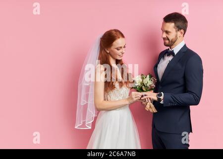 Handsome male in suit and female in white wedding dress putting golden ring with diamond on finger, stand isolated over pink background. Love, marriag Stock Photo