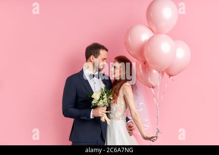 Charming lady with red hair and bearded man prepare to get married with each other, posing isolated over pink background with air balloons. Stock Photo