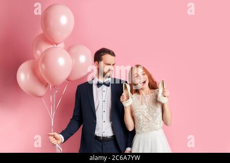 Serious male in suit holding pink air balloons look at woman in white wedding dress with opened mouth hold shoes isolated over pink background Stock Photo