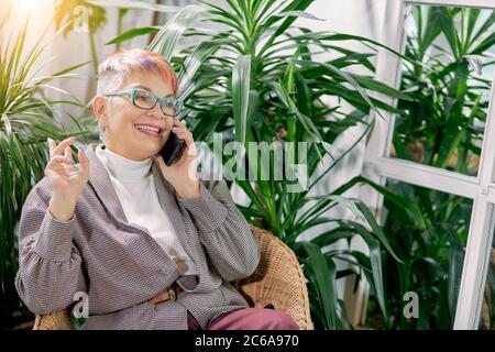 Actively gesturing senior businesswoman sitting on chair in business center full of green plants. Happy smile, call to friend Stock Photo